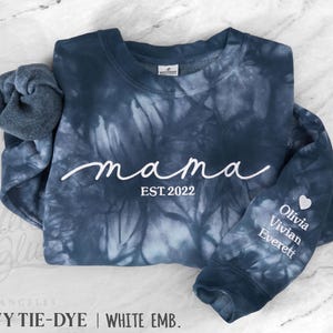 May include: A navy blue tie-dye sweatshirt featuring the white text mama EST. 2022 on the chest. The sleeve displays the names Olivia, Vivian, and Everett in white text beneath a small white heart icon. The fabric has a crew neckline.