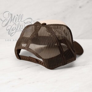 May include: A brown and tan trucker hat with a mesh back. The hat has a curved brim and an adjustable strap. The text "Mill Blue Los Angeles" is visible in the background.