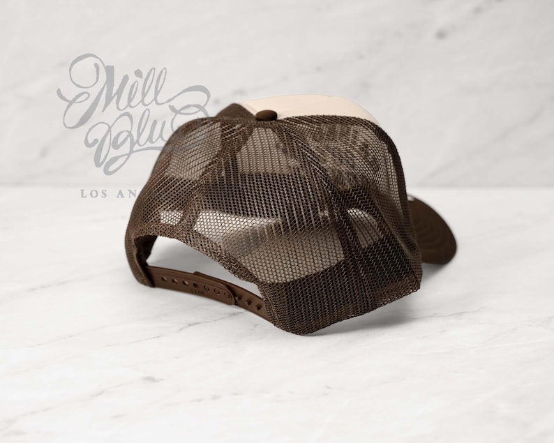 May include: A brown and tan trucker hat with a mesh back. The hat has a curved brim and an adjustable strap. The text "Mill Blue Los Angeles" is visible in the background.