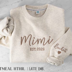 May include: An oatmeal heather crewneck sweatshirt featuring brown embroidery on the chest that reads Mimi EST. 2026. The sleeve is embroidered with the name Beckett and a small heart icon. The garment has a ribbed collar and cuffs.
