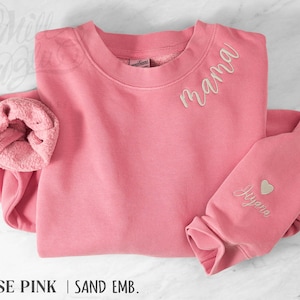 May include: A rose pink sweatshirt with the word "mama" embroidered in a cursive font on the chest. The sleeve features a heart and the name "Jayana" embroidered in a matching sand color. The sweatshirt is made of a soft material.