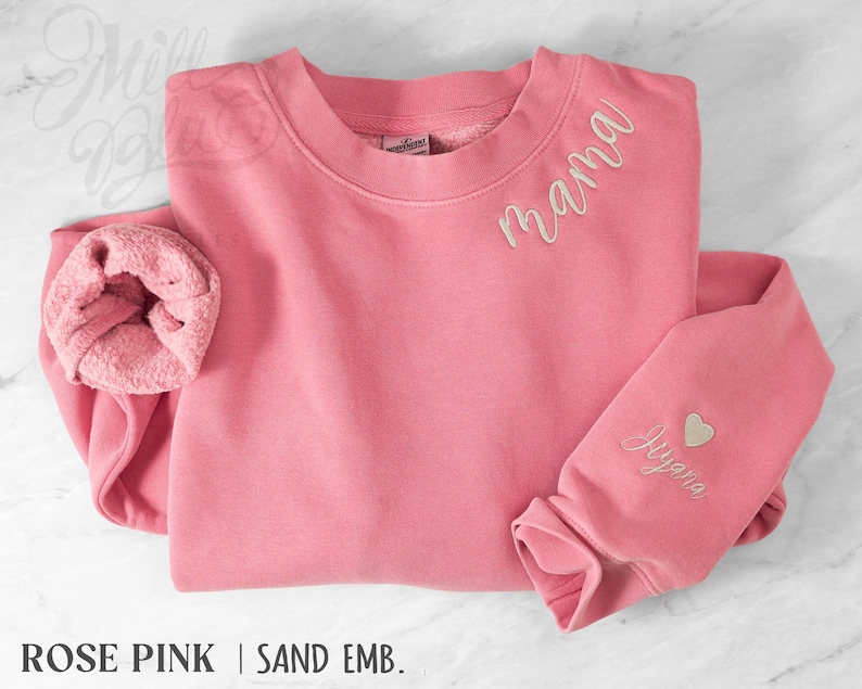 May include: A rose pink sweatshirt with the word "mama" embroidered in a cursive font on the chest. The sleeve features a heart and the name "Jayana" embroidered in a matching sand color. The sweatshirt is made of a soft material.