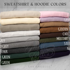 May include: A color chart titled SWEATSHIRT & HOODIE COLORS displaying folded fabric stacks in shades of sand, ivory, pebble, ash, light heather grey, oxford grey, dark heather, alpine green, forest green, light pink, lavender, camel, golden pecan, chocolate, military green, steel blue, navy heather, and navy.