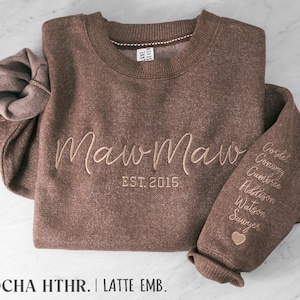 May include: A mocha heather brown crewneck sweatshirt featuring the embroidered text MawMaw EST. 2015 in latte-colored thread. The left sleeve is embroidered with a list of names including Cordet, Conway, Cambree, Addison, Watson, and Sawyer, finished with a small heart icon.