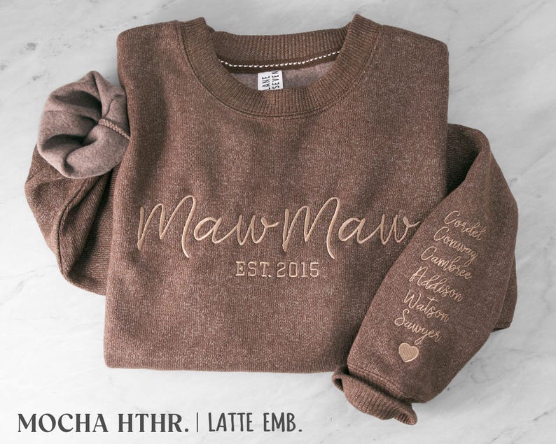 May include: A mocha heather brown crewneck sweatshirt featuring the embroidered text MawMaw EST. 2015 in latte-colored thread. The left sleeve is embroidered with a list of names including Cordet, Conway, Cambree, Addison, Watson, and Sawyer, finished with a small heart icon.