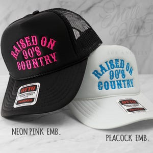 May include: Two trucker hats, one black and one white, with the embroidered text "RAISED ON 90'S COUNTRY." The black hat has neon pink embroidery, and the white hat has peacock blue embroidery. Both hats have a mesh back and a curved brim.