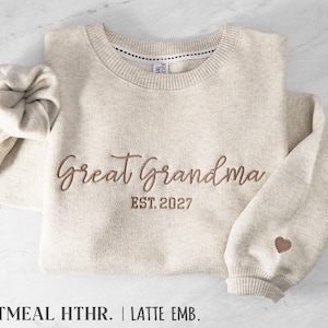 May include: An oatmeal heather crewneck sweatshirt featuring brown embroidery that reads Great Grandma EST. 2027 across the chest. A small brown embroidered heart is positioned on the left sleeve cuff. The garment has a ribbed neckline.
