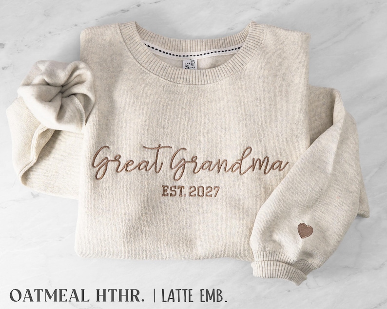 May include: An oatmeal heather crewneck sweatshirt featuring brown embroidery that reads Great Grandma EST. 2027 across the chest. A small brown embroidered heart is positioned on the left sleeve cuff. The garment has a ribbed neckline.