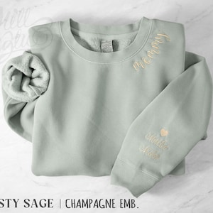 May include: A sage green sweatshirt with champagne-coloured embroidery. The word "Mommy" is embroidered on the chest, and a name and heart are on the sleeve. The text "Dusty Sage | Champagne Emb." is at the bottom.
