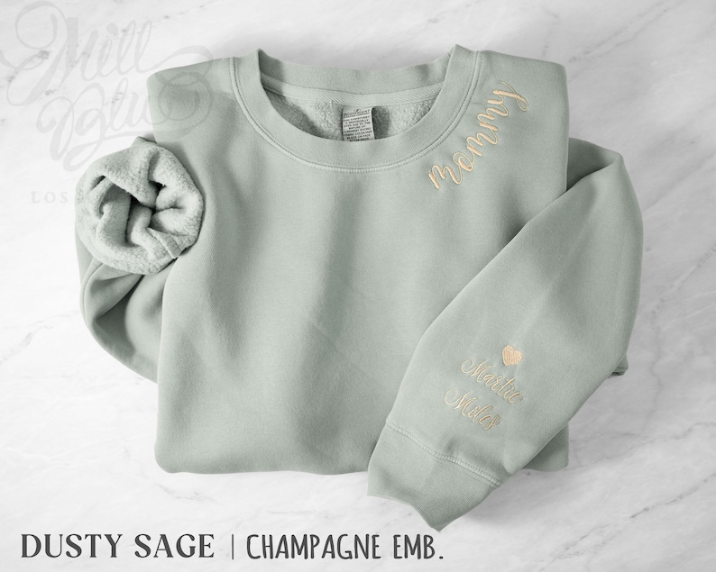 May include: A sage green sweatshirt with champagne-coloured embroidery. The word "Mommy" is embroidered on the chest, and a name and heart are on the sleeve. The text "Dusty Sage | Champagne Emb." is at the bottom.
