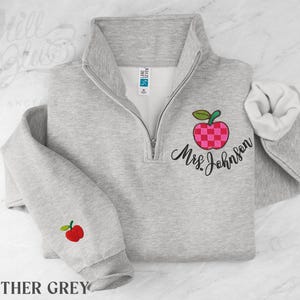 May include: A heather grey quarter-zip sweatshirt featuring an embroidered pink and red checkered apple design on the chest with the text Mrs. Johnson in black script. A smaller red apple is embroidered on the left sleeve cuff near the wrist.