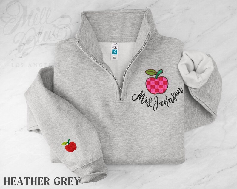 May include: A heather grey quarter-zip sweatshirt featuring an embroidered pink and red checkered apple design on the chest with the text Mrs. Johnson in black script. A smaller red apple is embroidered on the left sleeve cuff near the wrist.