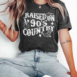 K&ouml;nnte beinhalten: Ein dunkelgraues T-Shirt mit dem Text "RAISED ON 90'S COUNTRY" in Wei&szlig;. Das Design beinhaltet eine Gitarre, einen Cowboyhut und einen Stiefel.