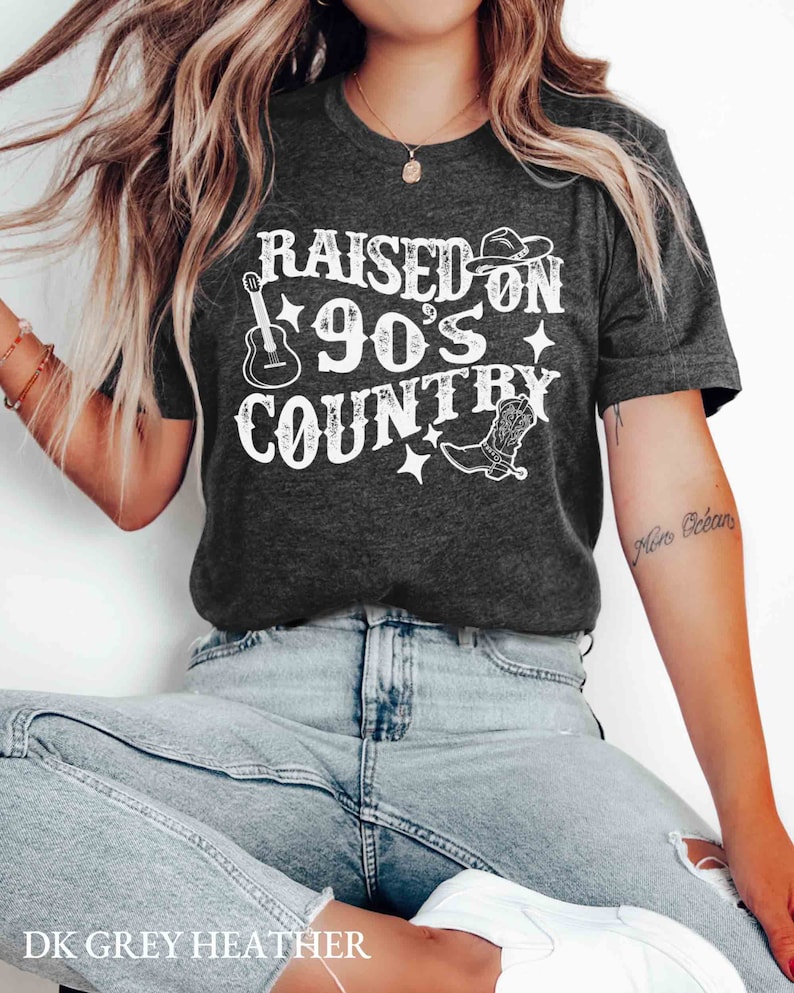 K&ouml;nnte beinhalten: Ein dunkelgraues T-Shirt mit dem Text "RAISED ON 90'S COUNTRY" in Wei&szlig;. Das Design beinhaltet eine Gitarre, einen Cowboyhut und einen Stiefel.