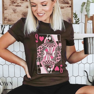 K&ouml;nnte beinhalten: Ein braunes T-Shirt mit einem pinkfarbenen und schwarzen Spielkartenmotiv der Herzdame. Das Motiv zeigt eine Frau mit Sonnenbrille und Krone, die ein Glas Wein h&auml;lt.