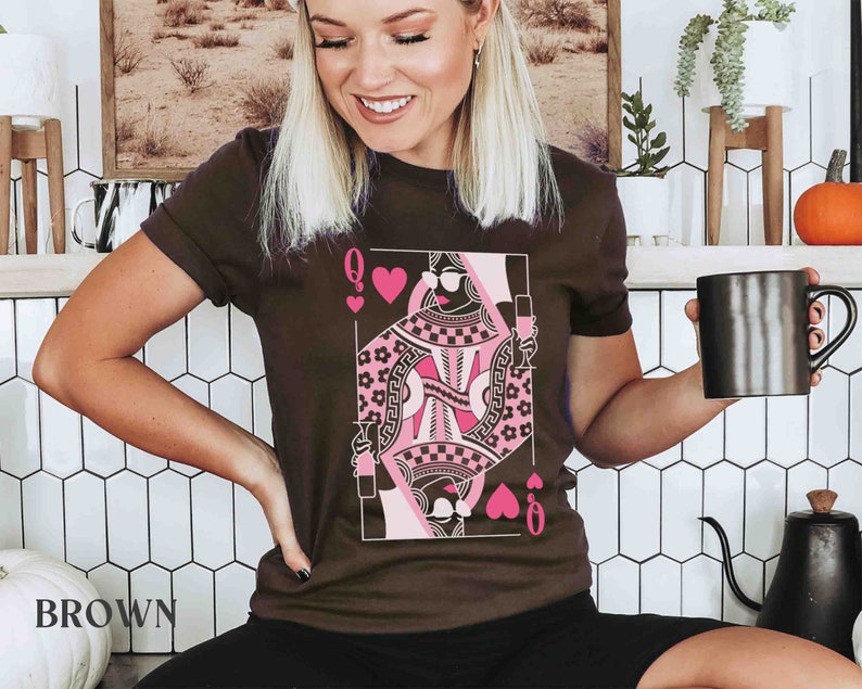 K&ouml;nnte beinhalten: Ein braunes T-Shirt mit einem pinkfarbenen und schwarzen Spielkartenmotiv der Herzdame. Das Motiv zeigt eine Frau mit Sonnenbrille und Krone, die ein Glas Wein h&auml;lt.