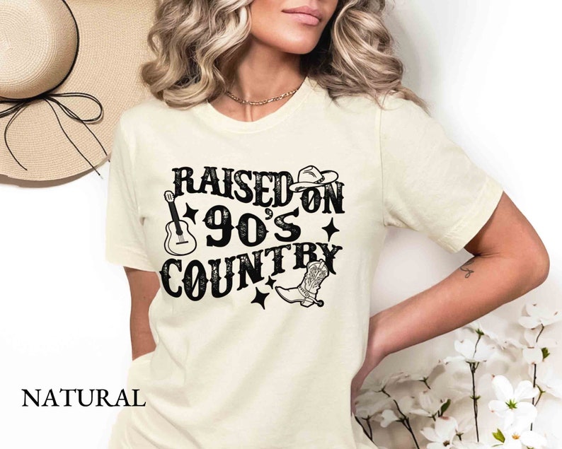 K&ouml;nnte beinhalten: Ein wei&szlig;es T-Shirt mit einem schwarzen Aufdruck, der "Raised on 90's Country" mit einer Gitarre, einem Cowboyhut und einem Stiefel zeigt.
