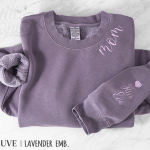 May include: Mauve sweatshirt with the word "mom" embroidered on the chest and names "Rico Eva" on the sleeve, plus a small pink heart. The sweatshirt has a fleecy interior and the text "MAUVE | LAVENDER EMB."