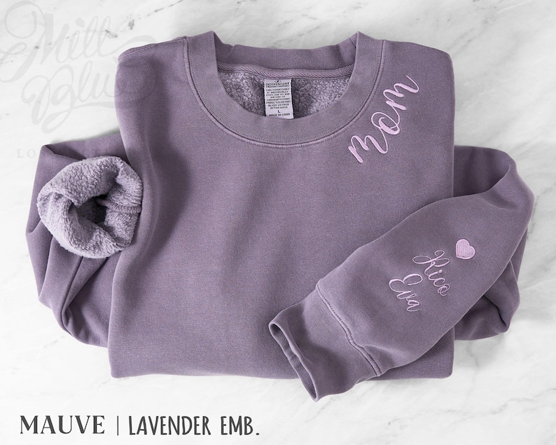 May include: Mauve sweatshirt with the word "mom" embroidered on the chest and names "Rico Eva" on the sleeve, plus a small pink heart. The sweatshirt has a fleecy interior and the text "MAUVE | LAVENDER EMB."