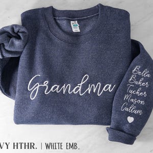 May include: A navy heather crewneck sweatshirt featuring the word Grandma embroidered in white script on the chest. The left sleeve displays a vertical list of names including Bella, Baker, Tucker, Mason, and Callum in white script, finished with a small white heart icon.
