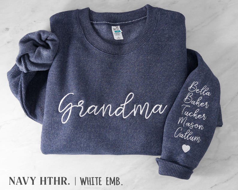 May include: A navy heather crewneck sweatshirt featuring the word Grandma embroidered in white script on the chest. The left sleeve displays a vertical list of names including Bella, Baker, Tucker, Mason, and Callum in white script, finished with a small white heart icon.
