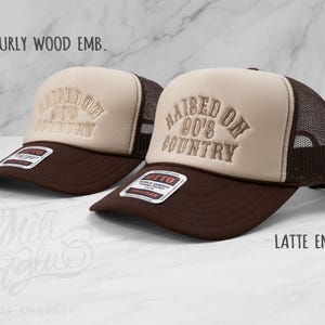 May include: Two brown and tan trucker hats with the embroidered text "RAISED ON 90'S COUNTRY." The hats have a mesh back and a curved brim. The text "BURLY WOOD EMB." and "LATTE EMB." are also visible.