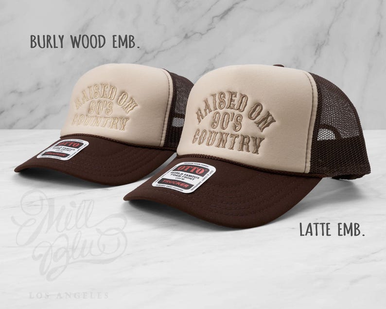 May include: Two brown and tan trucker hats with the embroidered text "RAISED ON 90'S COUNTRY." The hats have a mesh back and a curved brim. The text "BURLY WOOD EMB." and "LATTE EMB." are also visible.