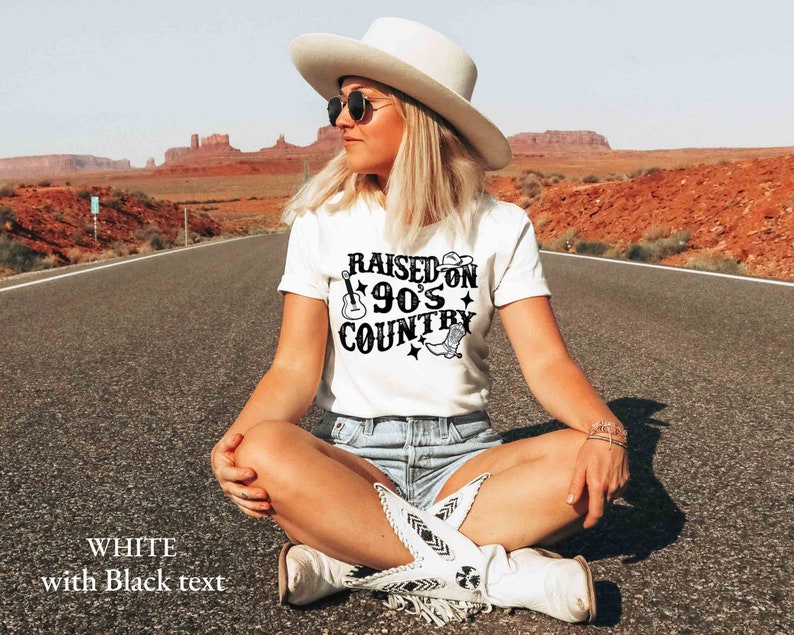 K&ouml;nnte beinhalten: Wei&szlig;es T-Shirt mit schwarzem Text, der "Raised on 90's Country" lautet. Der Text ist in einer Retro-Schriftart mit einem Gitarren- und Cowboystiefel-Motiv. Das Shirt wird von einer Person getragen, die auf einer Stra&szlig;e mit einer W&uuml;stenlandschaft im Hintergrund sitzt.