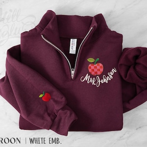 May include: A maroon quarter-zip sweatshirt featuring white embroidered text that reads Mrs. Johnson. The design includes a checkered red and pink apple graphic on the chest and a small solid red apple embroidered on the right sleeve cuff.