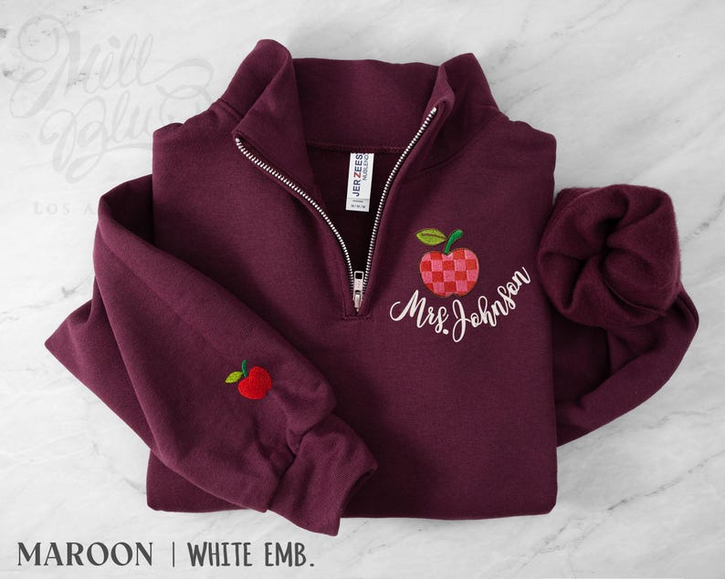 May include: A maroon quarter-zip sweatshirt featuring white embroidered text that reads Mrs. Johnson. The design includes a checkered red and pink apple graphic on the chest and a small solid red apple embroidered on the right sleeve cuff.