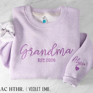 May include: A lilac heather crewneck sweatshirt featuring dark purple embroidery on the chest that reads Grandma EST. 2026. The sleeve is embroidered with the name Alyssa above a small purple heart. The interior has a white fleece lining.