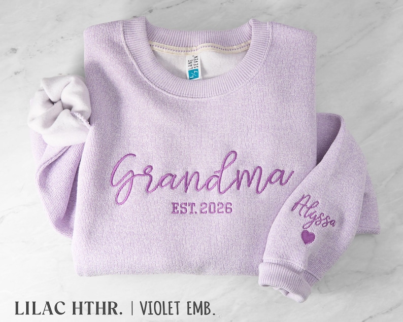 May include: A lilac heather crewneck sweatshirt featuring dark purple embroidery on the chest that reads Grandma EST. 2026. The sleeve is embroidered with the name Alyssa above a small purple heart. The interior has a white fleece lining.