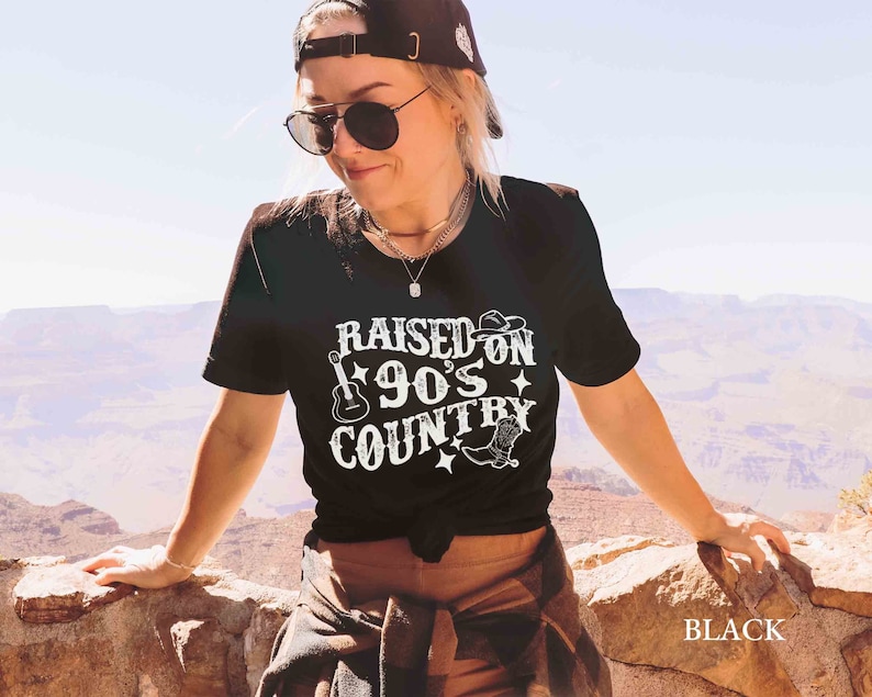 K&ouml;nnte beinhalten: Schwarzes T-Shirt mit wei&szlig;em Text "Raised on 90's Country" mit Gitarre und Cowboy-Stiefel-Grafik.