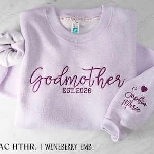 May include: A lilac crewneck sweatshirt featuring dark purple embroidery that reads Godmother EST. 2026 on the chest and Sophia Marie with a small heart on the sleeve. The garment has a ribbed collar and cuffs, with a Lane Seven brand tag inside.