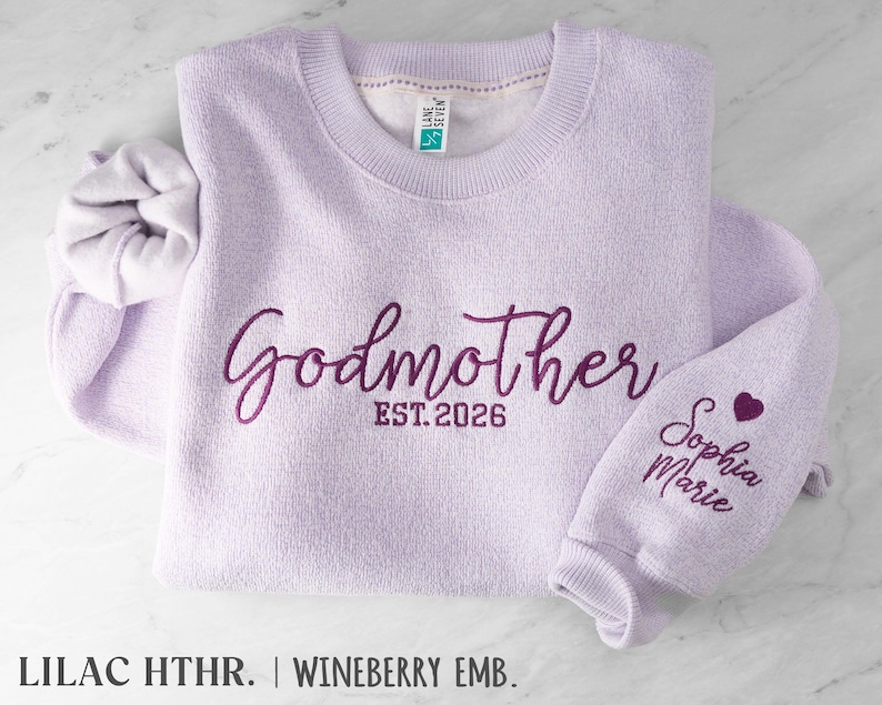 May include: A lilac crewneck sweatshirt featuring dark purple embroidery that reads Godmother EST. 2026 on the chest and Sophia Marie with a small heart on the sleeve. The garment has a ribbed collar and cuffs, with a Lane Seven brand tag inside.