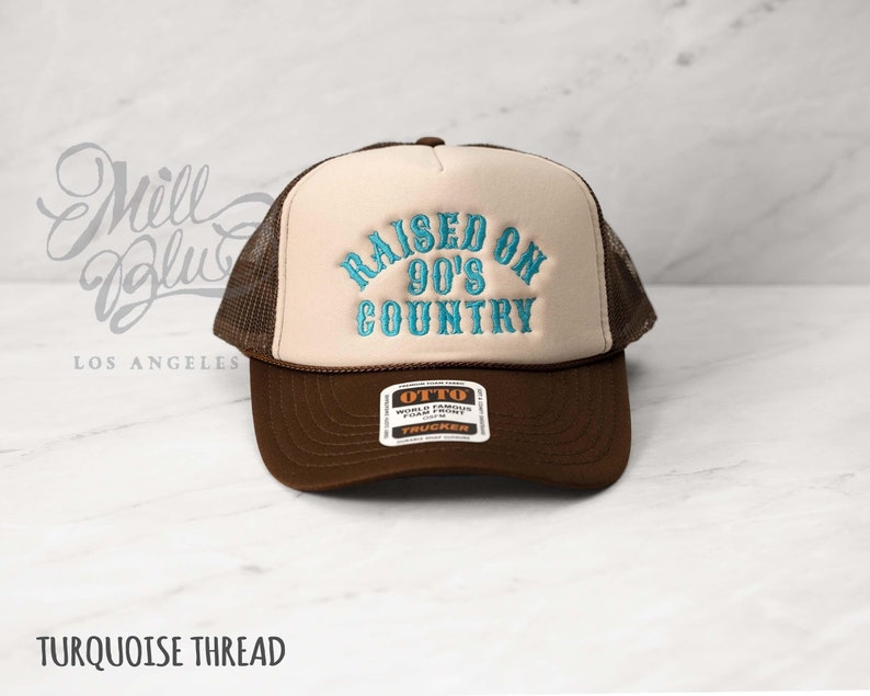 May include: A brown and tan trucker hat with the text "RAISED ON 90'S COUNTRY" embroidered in turquoise thread. The hat features a mesh back and a curved brim. The brand tag "OTTO" is visible on the front.