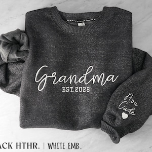 May include: A dark heather gray crewneck sweatshirt featuring white embroidery. The front displays the text "Grandma EST. 2026" in a script and block font. The sleeve is embroidered with the names "Ava" and "Cade" above a small heart.