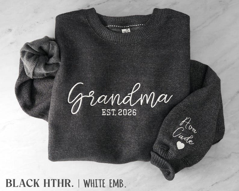 May include: A dark heather gray crewneck sweatshirt featuring white embroidery. The front displays the text "Grandma EST. 2026" in a script and block font. The sleeve is embroidered with the names "Ava" and "Cade" above a small heart.