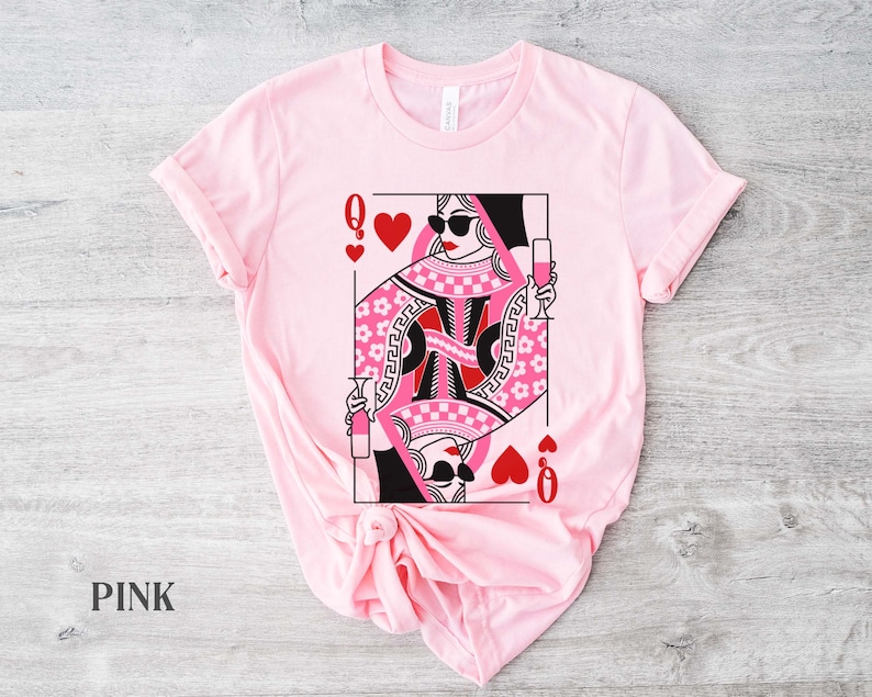 K&ouml;nnte beinhalten: Pinkes T-Shirt mit einem Grafikdruck einer Herzdame Spielkarte mit Sonnenbrille und einem Glas Champagner. Die Dame tr&auml;gt ein pinkfarbenes und schwarzes Karomuster.