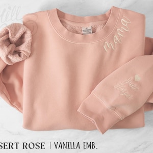 May include: A pale pink sweatshirt with the word "mama" embroidered in vanilla thread. The sleeve features the name "June Harper" and a heart. A matching scrunchie is next to the sweatshirt.