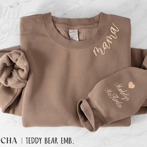 May include: A mocha-coloured sweatshirt with the word "mama" embroidered in a cursive font. The sleeve features the name "Madelyn McRenlee" and a heart. The sweatshirt has a teddy bear lining and the text "MOCHA | TEDDY BEAR EMB."