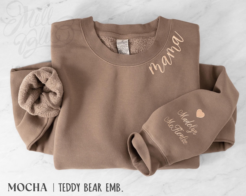 May include: A mocha-coloured sweatshirt with the word "mama" embroidered in a cursive font. The sleeve features the name "Madelyn McRenlee" and a heart. The sweatshirt has a teddy bear lining and the text "MOCHA | TEDDY BEAR EMB."