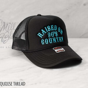 May include: Black trucker hat with a mesh back and a curved brim. The hat features the embroidered text "RAISED ON 90'S COUNTRY" in turquoise thread. The hat has a black rope detail and a snap closure.