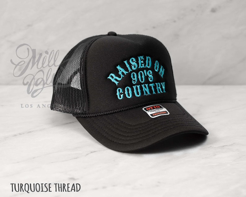 May include: Black trucker hat with a mesh back and a curved brim. The hat features the embroidered text "RAISED ON 90'S COUNTRY" in turquoise thread. The hat has a black rope detail and a snap closure.