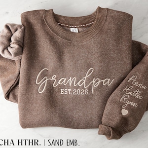May include: A mocha heather brown crewneck sweatshirt featuring cream-colored embroidery that reads "Grandpa EST. 2026" on the chest. The sleeve is embroidered with the names "Anna," "Hallie," and "Ryan" above a small heart icon.