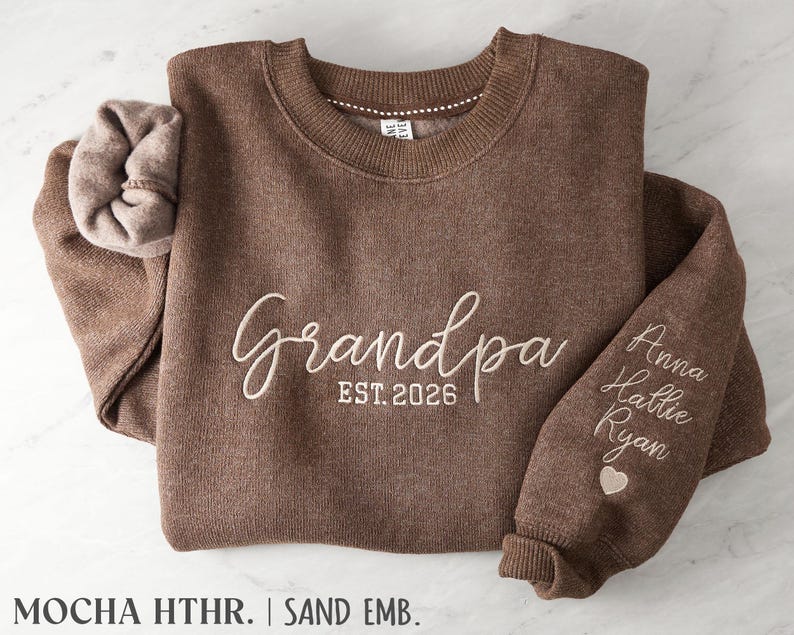 May include: A mocha heather brown crewneck sweatshirt featuring cream-colored embroidery that reads "Grandpa EST. 2026" on the chest. The sleeve is embroidered with the names "Anna," "Hallie," and "Ryan" above a small heart icon.