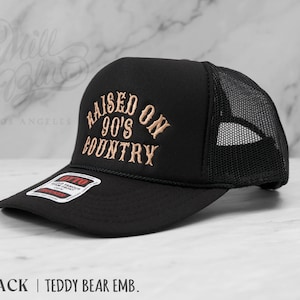 May include: Black trucker hat with a mesh back and a curved brim. The hat features the embroidered text "RAISED ON 90'S COUNTRY" in a tan color. The hat also has a small tag that reads "OTTO".