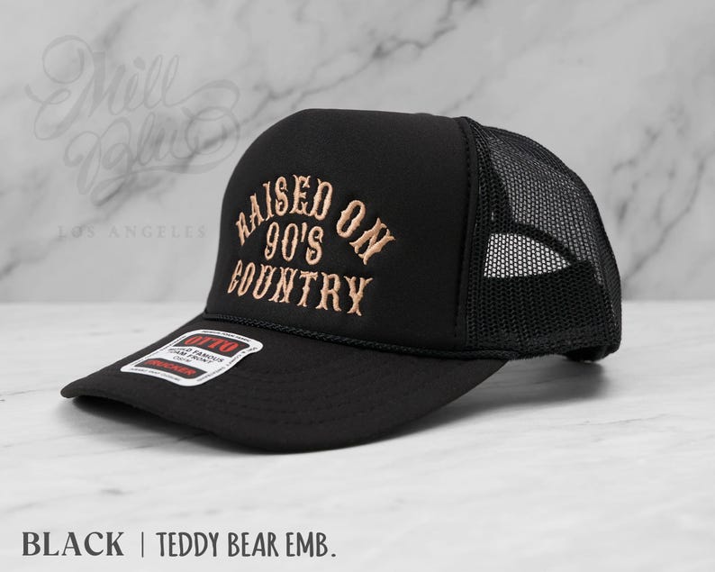 May include: Black trucker hat with a mesh back and a curved brim. The hat features the embroidered text "RAISED ON 90'S COUNTRY" in a tan color. The hat also has a small tag that reads "OTTO".