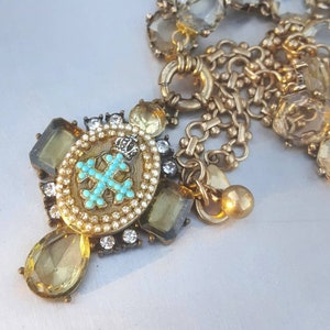 Peut inclure: Un bracelet à breloques en ton or avec un grand pendentif orné d'une croix turquoise et d'une couronne. Le pendentif est entouré de cristaux facetés clairs et jaunes.