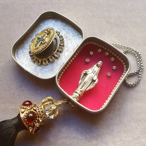 May include: A silver charm necklace with a small, silver statue of the Virgin Mary in a red velvet box. The box is decorated with white beads and rhinestones. The necklace has a black tassel with a gold crown and red gems.